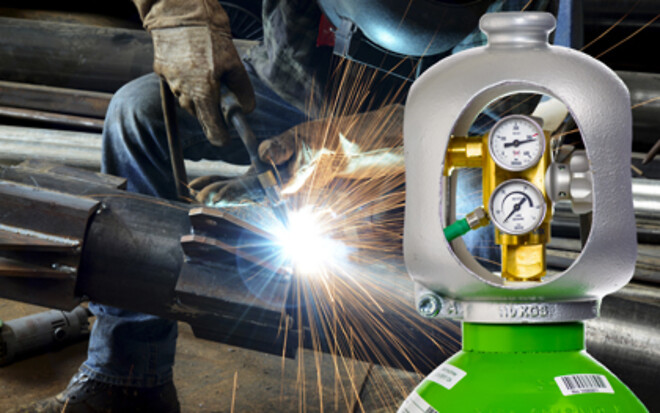 Gas cylinder with pressure regulators in the foreground, positioned in front of a metal welding operation producing bright sparks.