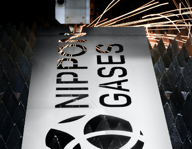 Laser cutting machine processing a metal sheet with the Nippon Gases logo and text, producing bright sparks.