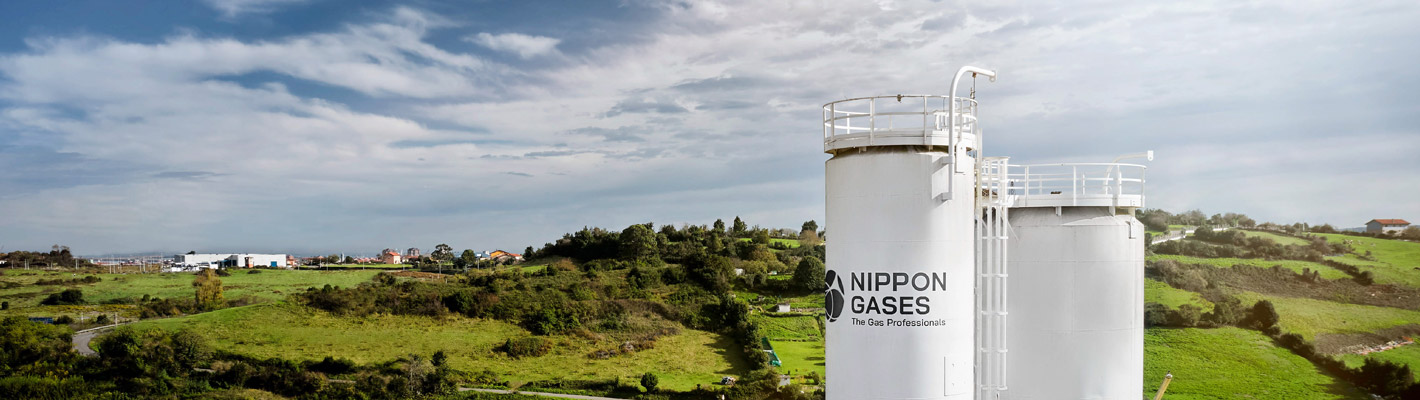 Large industrial gas storage tanks branded “Nippon Gases” in a green rural landscape under a cloudy sky.