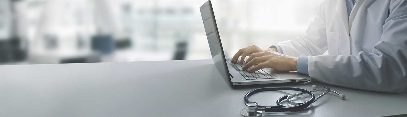 Close-up of a healthcare professional using a laptop in a clinical setting, with a stethoscope resting on a desk in the foreground.