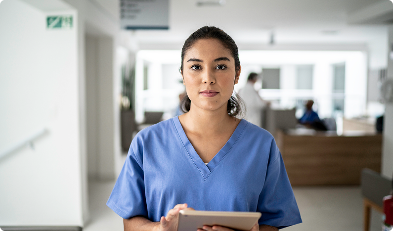 Healthcare professional in blue scrubs holding a tablet in a hospital corridor, with a reception area blurred in the background.