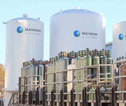 Industrial gas cylinder storage area with rows of compressed gas cylinders in front of large Matheson bulk gas tanks.